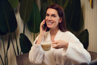 美丽的女人持有杯卡布奇诺咖啡微笑相机美丽的露出牙齿的会说话的移动电话放松水疗中心休息室接收健康美治疗
