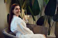 迷人的欧洲中间岁的女人白色浴袍享受咖啡打破坐着扶手椅休息室现代水疗中心诊所微笑高高兴兴地相机