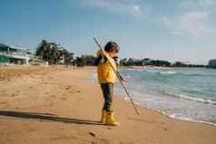 男孩黄色的橡胶靴子玩坚持沙子海滩学校孩子触碰水秋天冬天海孩子有趣的波海岸春天假期假期概念