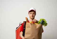 快乐交付男人。红色的统一的帽热袋背包持有棕色（的）工艺纸袋食物产品孤立的灰色的背景工作室交付服务商店餐厅首页