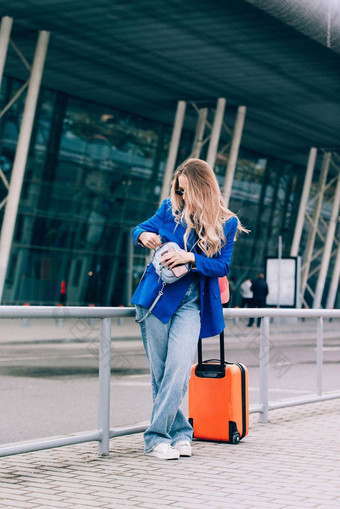 肖像旅行者女人站橙色手提箱机场年轻的时尚女人蓝色的牛仔裤夹克黑色的衬衫白色运动鞋
