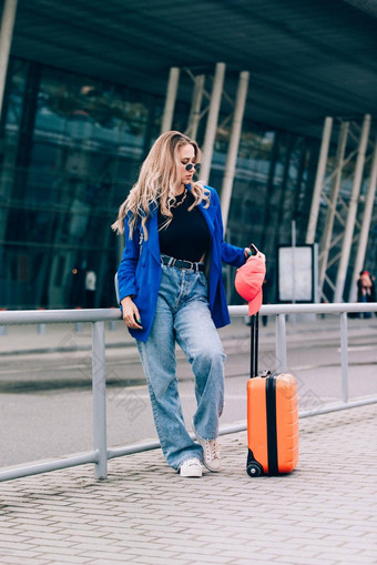 肖像旅行者女人站橙色手提箱机场年轻的时尚女人蓝色的牛仔裤<strong>夹克</strong>黑色的衬衫白色<strong>运动</strong>鞋