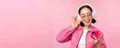 时尚的亚洲女孩跳舞智能手机听音乐耳机移动电话应用程序微笑笑摆姿势粉红色的背景