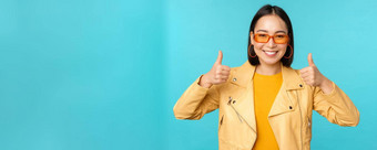 肖像现代年轻的亚洲中国人女人太阳镜穿时尚的外套显示拇指微笑很高兴推荐smth蓝色的背景