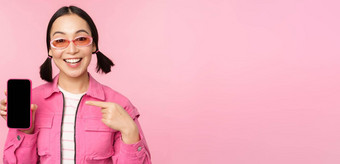 <strong>热情</strong>的亚洲女人时尚的衣服太阳镜指出手指移动电话屏幕显示智能手机应用程序站粉红色的<strong>背景</strong>