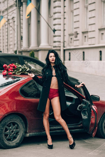 优雅的美丽的浅黑肤色的女人女人红色的短裤黑色的外套摆姿势红色的车大花束红色的玫瑰车屋顶