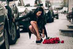 优雅的美丽的浅黑肤色的女人女人红色的短裤黑色的外套摆姿势城市街道大花束红色的玫瑰生日