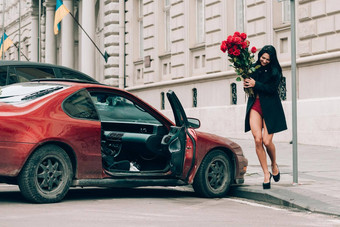 优雅的美丽的浅黑肤色的女人女人红色的短裤黑色的外套摆姿势红色的车大花束红色的玫瑰生日3月情人节一天