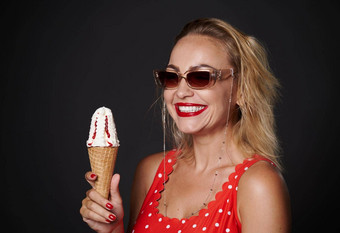 快乐的中间岁的金发女郎女人穿保护太阳镜红色的泳衣持有冰淇淋锥<strong>微笑</strong>露出牙齿的<strong>微笑</strong>相机孤立的黑色的背景复制空间