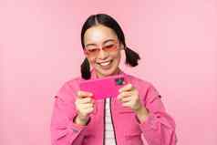 肖像快乐亚洲女孩玩智能手机看视频移动电话应用程序站粉红色的背景