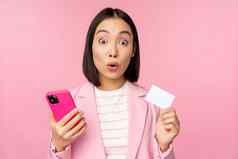 惊讶女商人亚洲女孩西装显示信贷卡移动电话订单在线购物智能手机站粉红色的背景广告概念