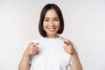 图像微笑亚洲女孩指出手指相机选择邀请祝贺站T恤白色背景