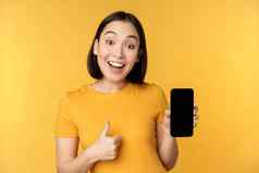 兴奋亚洲女人显示移动电话屏幕拇指smth好推荐智能手机应用程序站黄色的背景