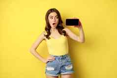 震惊了惊讶时尚的女孩显示智能手机屏幕应用程序水平移动电话显示站夏天衣服黄色的背景