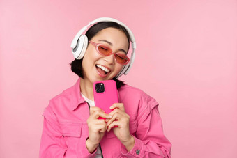 时尚的亚洲女孩耳机听音乐采取照片移动电话智能手机站粉红色的背景