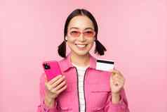 在线购物微笑亚洲女孩购物者持有智能手机信贷卡支付移动应用程序站粉红色的背景