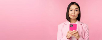 怀疑女商人西装持有智能手机<strong>扮鬼</strong>脸使持怀疑态度脸表达式站粉红色的背景