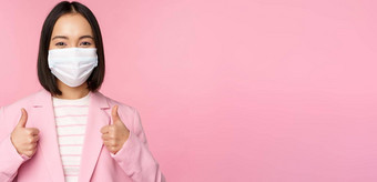 亚洲女商人西装医疗脸面具显示拇指推荐穿个人保护设备办公室科维德流感大流行粉红色的背景