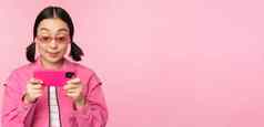 肖像快乐亚洲女孩玩智能手机看视频移动电话应用程序站粉红色的背景