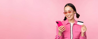 在线购物微笑亚洲女孩购物者持有智能<strong>手机</strong>信贷卡支付移动应用程序站粉红色的背景