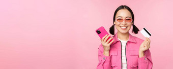 美丽的朝鲜文女人持有智能<strong>手机</strong>信贷卡微笑相机购买在线购物移动电话站粉红色的背景