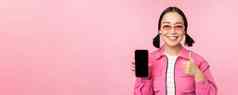快乐时尚的女孩建议应用程序移动电话微笑亚洲女模型显示智能手机屏幕拇指站粉红色的背景