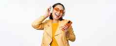 技术概念时尚的亚洲女孩耳机持有智能手机跳舞唱歌听音乐站白色背景