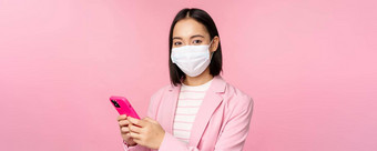 亚洲女商人医疗脸面具移动电话日本语女售货员企业夫人西装持有智能手机站粉红色的背景