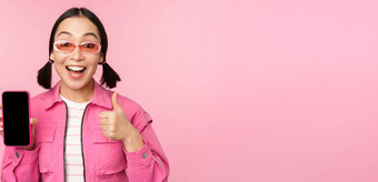 快乐时尚的女孩建议应用程序移动电话微笑亚洲女模型显示智能<strong>手机</strong>屏幕拇指站粉红色的背景