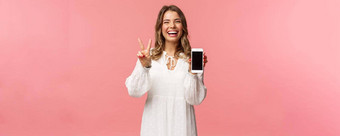 肖像卡哇伊乐观快乐年轻的女孩白色衣服显示移动电话显示和平标志笑感觉快乐的很高兴<strong>分享</strong>太棒了应用程序<strong>链接</strong>粉红色的背景