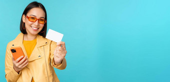 在线购物时尚的年轻的亚洲女人太阳镜显示信贷卡智能手机支付互联网使购买站蓝色的背景