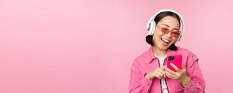 时尚的亚洲女孩跳舞智能<strong>手机</strong>听音乐耳机移动电话应用程序微笑笑摆姿势粉红色的背景
