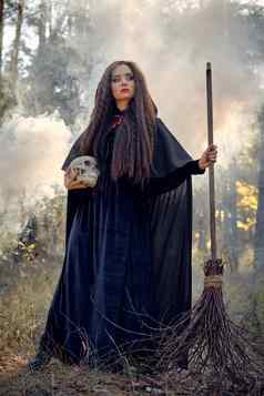 女巫黑色的长衣服角罩摆姿势烟松森林持有头骨扫帚法术魔法巫术完整的长度