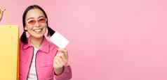 快乐年轻的亚洲女人显示信贷卡购物持有袋购买出售商店商店站粉红色的背景