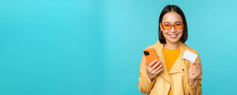 在线购物时尚的年轻的亚洲女人太阳镜显示信贷卡智能手机支付互联网使购买站蓝色的背景