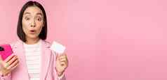 惊讶女商人亚洲女孩西装显示信贷卡移动电话订单在线购物智能手机站粉红色的背景广告概念