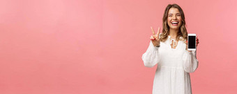 肖像卡哇伊乐观快乐年轻的女孩白色衣服显示移动电话显示和平标志笑感觉快乐的很高兴分享太棒了应用程序链接粉红色的背景