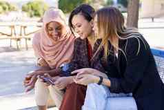 积极的多民族女朋友分享智能手机休息板凳上公园