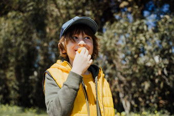 微笑快乐的孩子孩子黄色的背心绿色连帽衫吃脆零食在户外公共公园小学生男孩享受消耗咀嚼垃圾食物树植被背景