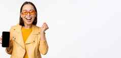 快乐亚洲女孩太阳镜显示移动电话屏幕智能手机接口笑微笑庆祝站白色背景
