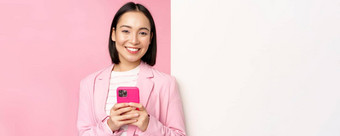 图像朝鲜文女企业家西装站信息墙广告董事会持有智能手机微笑摆姿势粉红色的背景