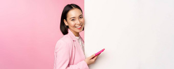 图像朝鲜文女企业家西装站信息墙广告董事会持有智能手机微笑摆姿势粉红色的背景