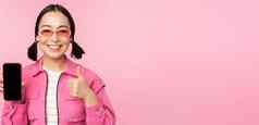 快乐时尚的女孩建议应用程序移动电话微笑亚洲女模型显示智能手机屏幕拇指站粉红色的背景