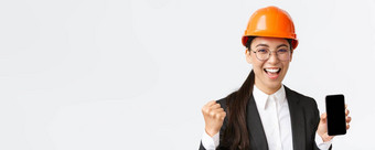 成功的赢得亚洲女工程师架构师安全头盔西装显示智能手机屏幕拳头泵大喊大叫喜乐并成就白色背景