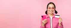 在线购物微笑亚洲女孩购物者持有智能手机信贷卡支付移动应用程序站粉红色的背景