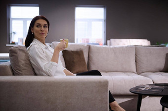 放松美丽的欧洲女人喝咖啡坐着舒适的沙发家具商店展厅