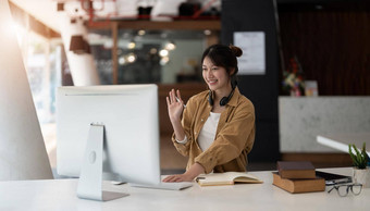 快乐的年轻的亚洲女无线耳机挥舞着魔杖问候屏幕坐着表格视频闲谈，聊天业务合作伙伴移动PC舒适的办公室