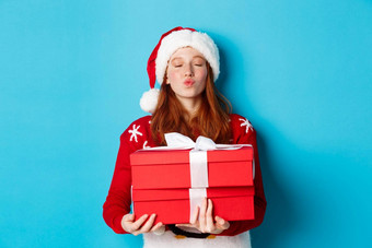 快乐假期圣诞节概念可爱的红色头发的人女孩持有礼物皱纹嘴唇吻穿圣诞老人他有趣的毛衣蓝色的背景