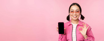 热情的年轻的亚洲女人显示标志微笑很高兴移动电话屏幕智能<strong>手机</strong>应用程序站粉红色的背景