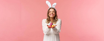 假期春天聚会，派对概念肖像兴奋迷人的金发碧眼的女人可爱的很高兴微笑持有画鸡蛋穿复活节一天装兔子耳朵白色衣服粉红色的背景